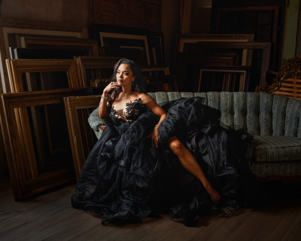 Elegant woman in a black lace gown lounging on a vintage patterned sofa in a dimly lit room filled with stacked ornate picture frames behind her.