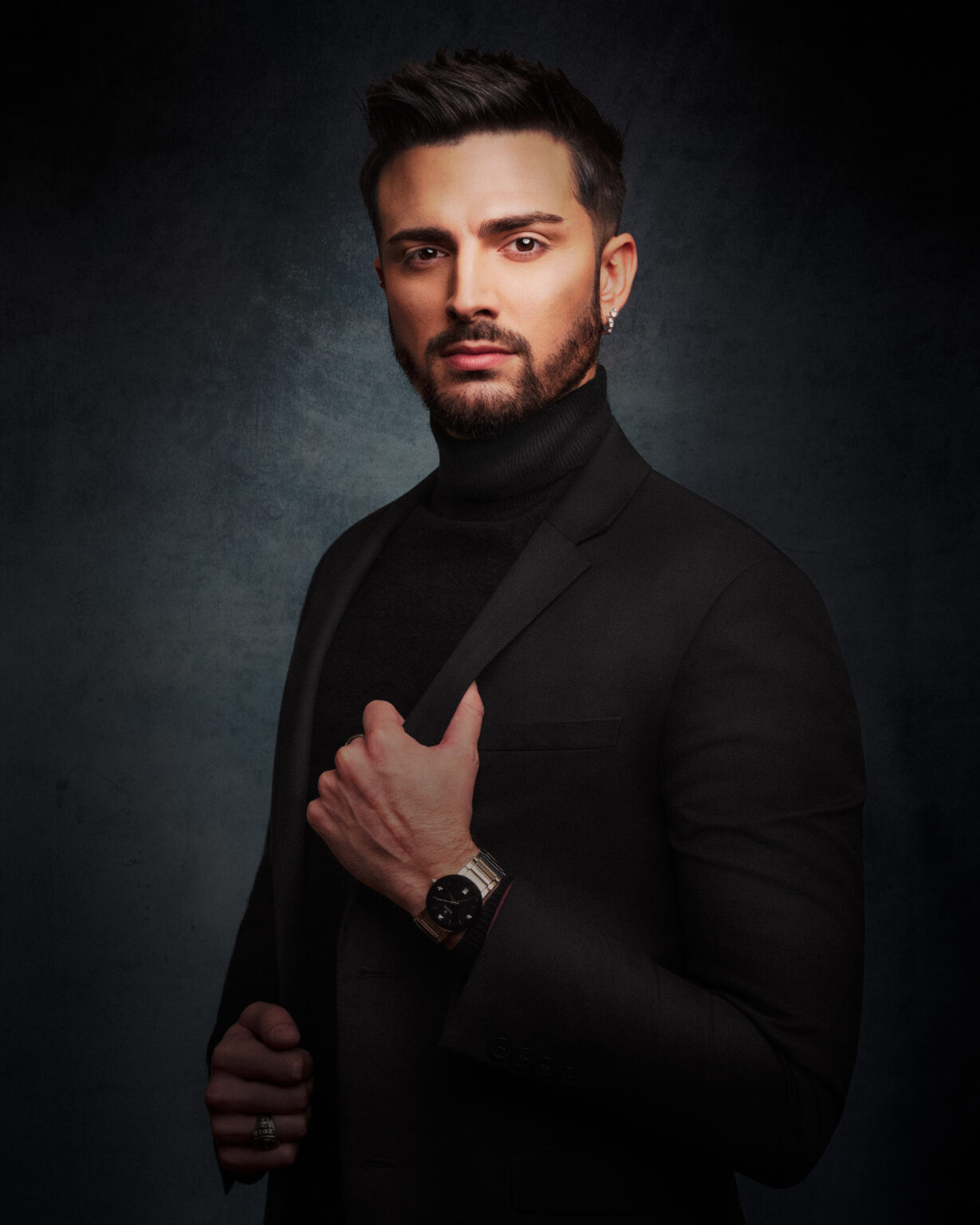 Male portrait in a black turtleneck and blazer, looking confidently at the camera against a dark textured background.