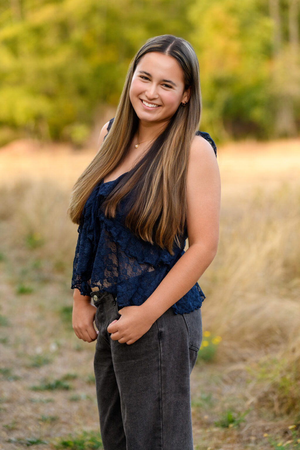Smiling young woman with long hair in a navy lace top standing in a sunlit field with a blurred yellow-green background.