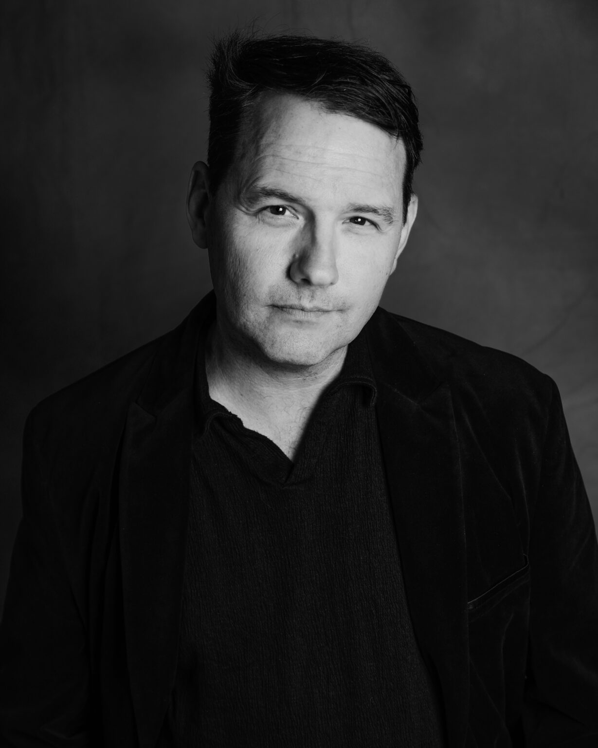Black-and-white studio portrait of a man in a dark jacket, facing the camera with a subtle smile against a dark backdrop.
